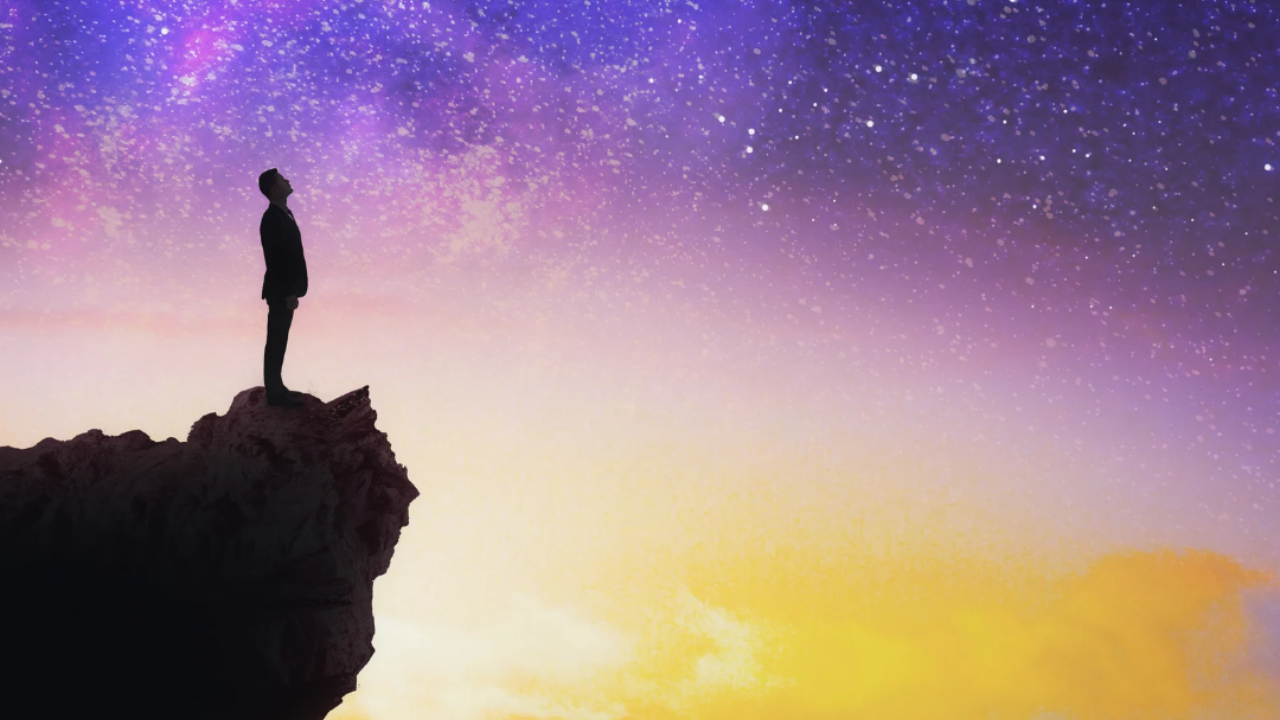 A man in silhouette stands at the edge of a rocky cliff, looking up into a purple starry night sky that fades to a golden glow.